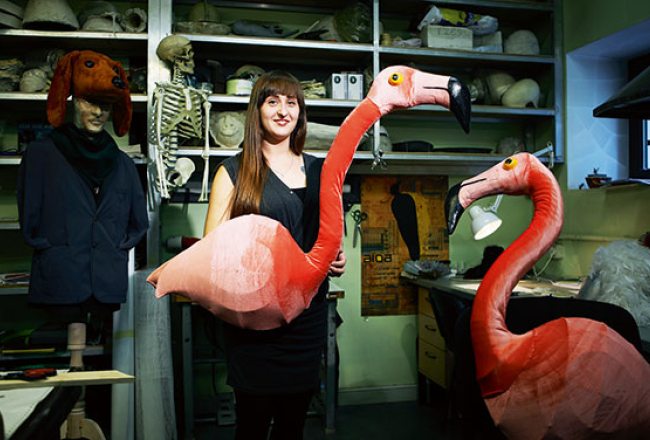 Latvijas Nacionālās operas un baleta butafore Ilze Egle ar flamingo maskām, kas liekamas nevis uz sejas, bet uz rokām. Ilzei aiz muguras plauktos glabājas visu masku atlējumi ģipsī vai mālā.