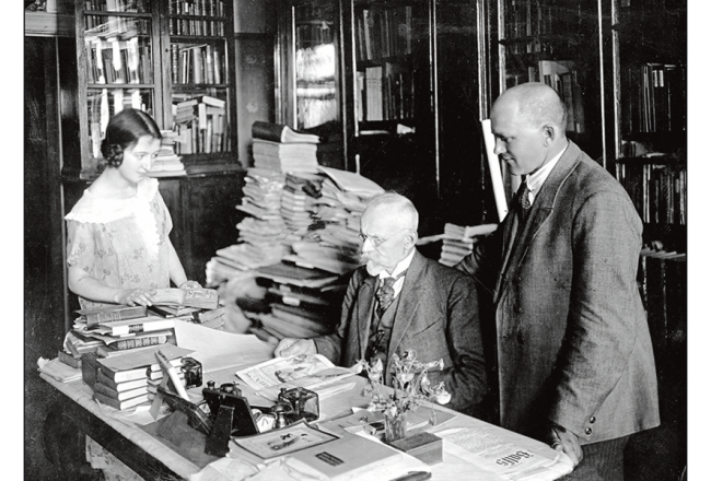 Jānis Misiņš (vidū) ar audžumeitu Hermīni un bibliofilu Fišeru savā mājas bibliotēkā Rīgā. 20. gadsimta 20. gadi. Fotogrāfs nezināms. LU bibliotēkas Misiņa bibliotēkas krājums