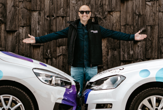 Edgars Bērziņš, autoskolas "Einšteins" vadītājs, stāv starp divām mācību automašīnām ar logo uz tumša koka fona, smaidot.