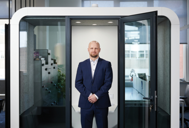 Vīrietis tumši zilā uzvalkā stāv stiklotā biroja kabīnē ar atvērtām durvīm, fonā redzams birojs.