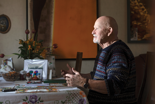 Pēteris Vasks, sirmgalvis ar bārdu, sēž pie galda un žestikulē, stāstot par saviem skaņdarbiem. Viņam ir tumši zils rakstains džemperis.