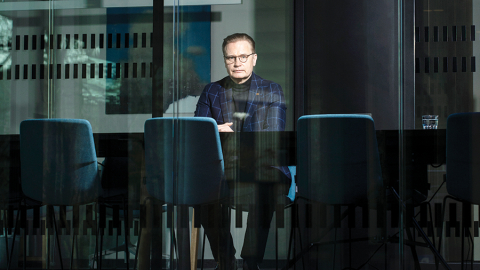 Uldis Mauriņš sēž pie galda stiklotā birojā, ar rokām sakrustotām priekšā. Viņš ir ģērbies uzvalkā un brillēs.