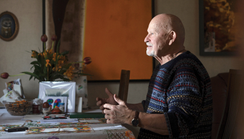 Pēteris Vasks, sirmgalvis ar bārdu, sēž pie galda un žestikulē, stāstot par saviem skaņdarbiem. Viņam ir tumši zils rakstains džemperis.
