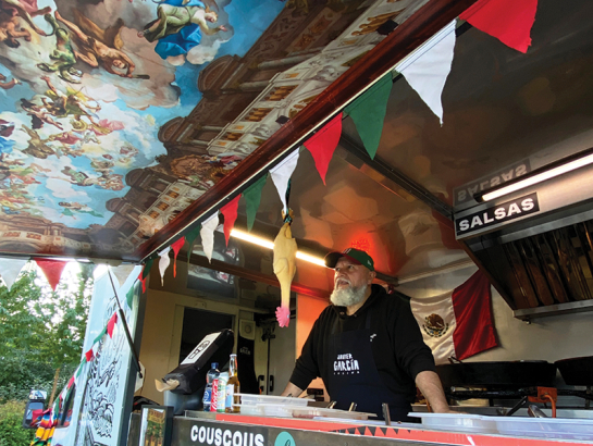 Food truck interjers ar Havjeru Garsiju priekšplānā, valkājot priekšautu ar vārdu "Javier Garcia Cocina". Pie griestiem piekārts gumijas cālis. Sānu durvis pārtapušas par nojumi ar fresku gleznojumu.