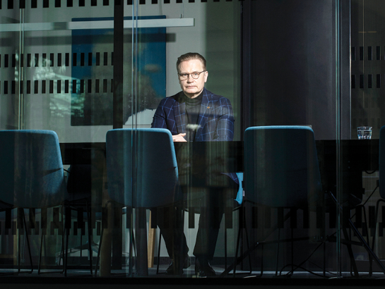 Uldis Mauriņš sēž pie galda stiklotā birojā, ar rokām sakrustotām priekšā. Viņš ir ģērbies uzvalkā un brillēs.