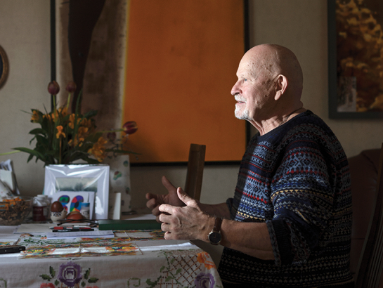 Pēteris Vasks, sirmgalvis ar bārdu, sēž pie galda un žestikulē, stāstot par saviem skaņdarbiem. Viņam ir tumši zils rakstains džemperis.