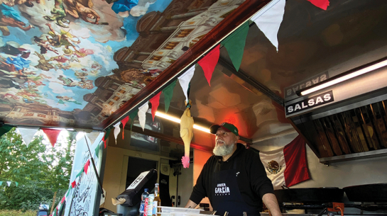 Food truck interjers ar Havjeru Garsiju priekšplānā, valkājot priekšautu ar vārdu "Javier Garcia Cocina". Pie griestiem piekārts gumijas cālis. Sānu durvis pārtapušas par nojumi ar fresku gleznojumu.