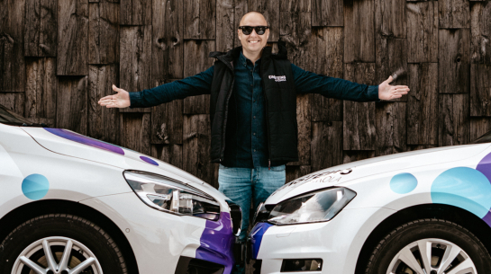 Edgars Bērziņš, autoskolas "Einšteins" vadītājs, stāv starp divām mācību automašīnām ar logo uz tumša koka fona, smaidot.