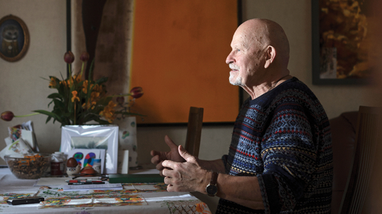 Pēteris Vasks, sirmgalvis ar bārdu, sēž pie galda un žestikulē, stāstot par saviem skaņdarbiem. Viņam ir tumši zils rakstains džemperis.