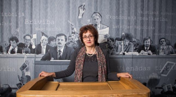 LTF muzeja vadītāja Meldra Usenko pie vēsturiskās Kongresu nama tribīnes. Viņai aizmugurē palielināta fotogrāfija no LTF dibināšanas kongresa prezidija, kas atradās uz Kongresu nama skatuves. Foto: Andrejs Terentjevs, F64