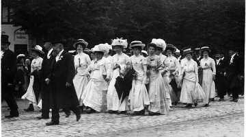 Rīga, 1910. gada 18. jūnijs. Rīgas latviešu pilsonības dāmas un kungi V Vispārējo latviešu dziesmu svētku gājienā. Svētki notika neapbūvētā teritorijā tagadējās Emiļa Melngaiļa un Hanzas ielas rajonā. 
Fotogrāfs nezināms. Latvijas Nacionālās bibliotēkas krājums