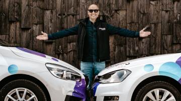 Edgars Bērziņš, autoskolas "Einšteins" vadītājs, stāv starp divām mācību automašīnām ar logo uz tumša koka fona, smaidot.