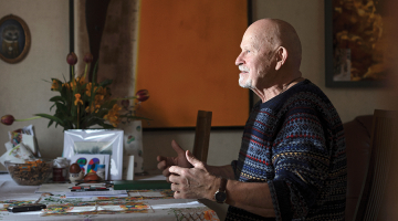 Pēteris Vasks, sirmgalvis ar bārdu, sēž pie galda un žestikulē, stāstot par saviem skaņdarbiem. Viņam ir tumši zils rakstains džemperis.