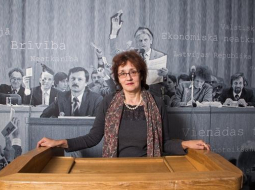 LTF muzeja vadītāja Meldra Usenko pie vēsturiskās Kongresu nama tribīnes. Viņai aizmugurē palielināta fotogrāfija no LTF dibināšanas kongresa prezidija, kas atradās uz Kongresu nama skatuves. Foto: Andrejs Terentjevs, F64