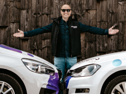 Edgars Bērziņš, autoskolas "Einšteins" vadītājs, stāv starp divām mācību automašīnām ar logo uz tumša koka fona, smaidot.